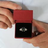 A man opening a ring box revealing a unique unheated 6.06ct teal sapphire, elongated cushion cut centre stone, with 4  tapering diamond oval side stones. Designed by engagement ring designer Antonia Guise.