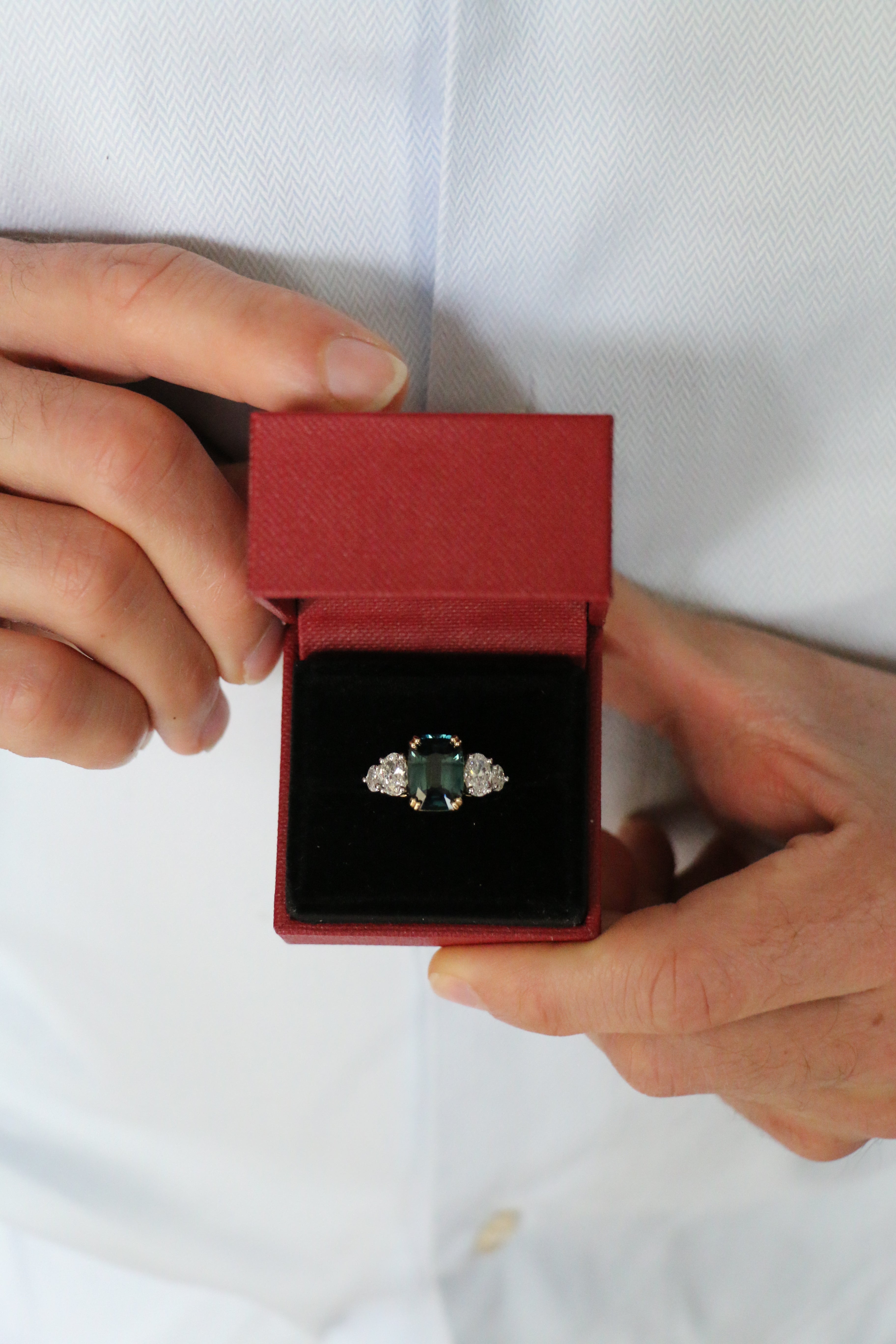 A man opening a ring box revealing a unique unheated 6.06ct teal sapphire, elongated cushion cut centre stone, with 4  tapering diamond oval side stones. Designed by engagement ring designer Antonia Guise.