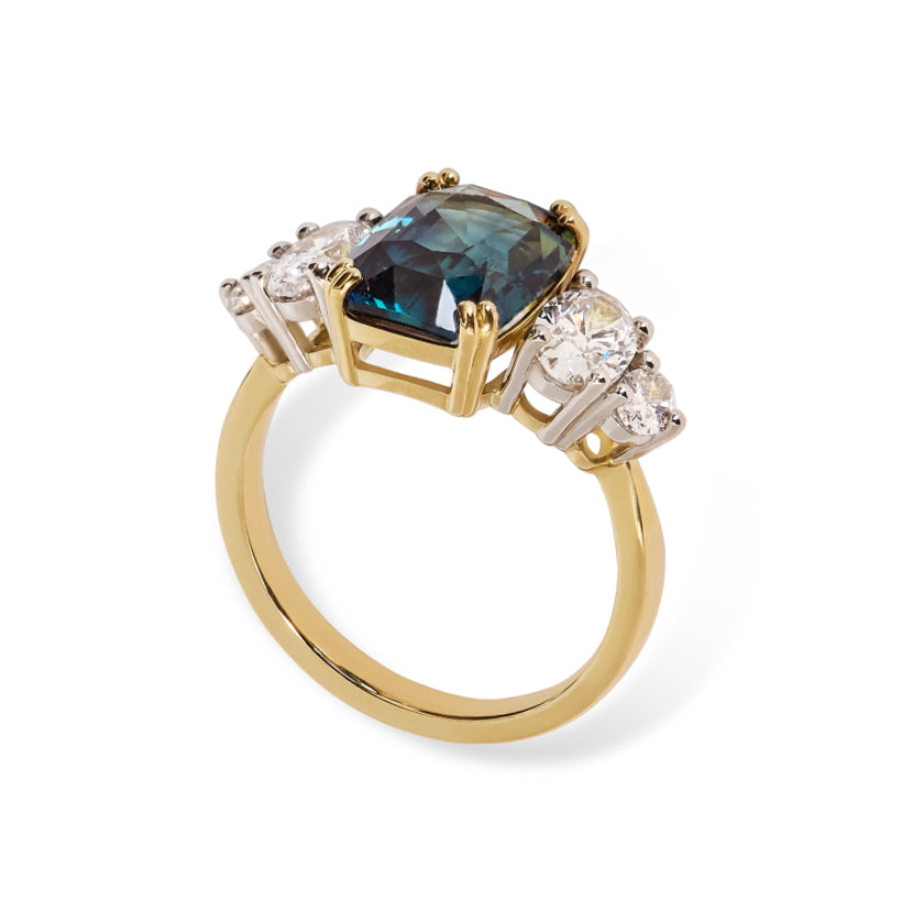 A  3/4 view of a unique unheated 6.06ct teal sapphire, elongated cushion cut centre stone, with 4  tapering diamond oval side stones. All set in recycled gold and platinum. Designed by London based engagement ring designer Antonia Guise. On a white background.